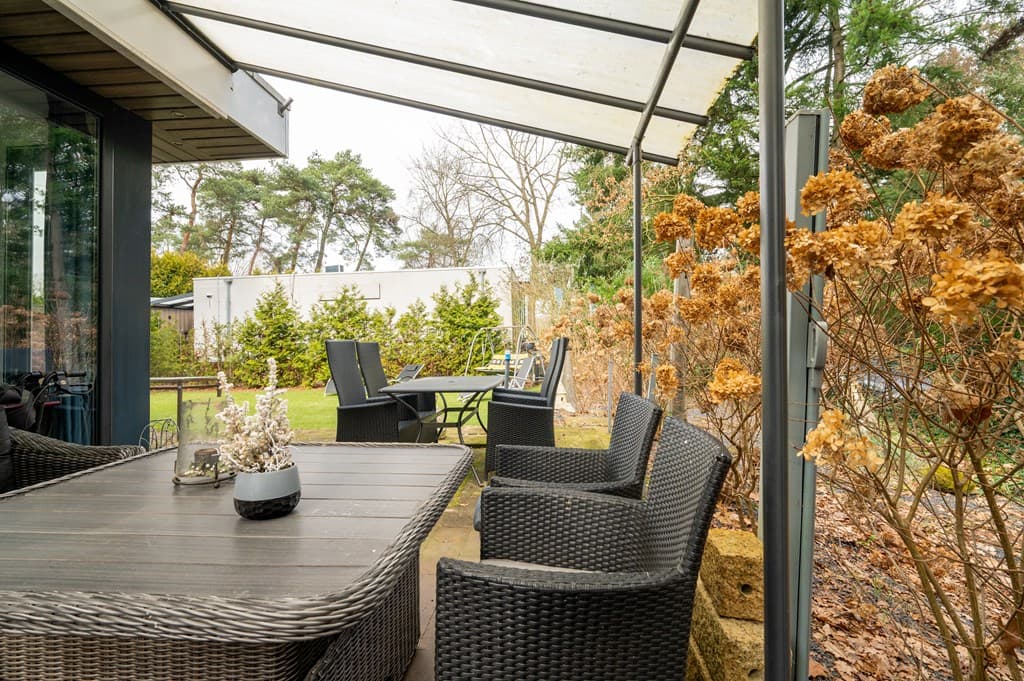Terras met een houten tafel en stoelen, omringd door droge planten en zicht op een groene tuin bij Camping De Konijnenberg.