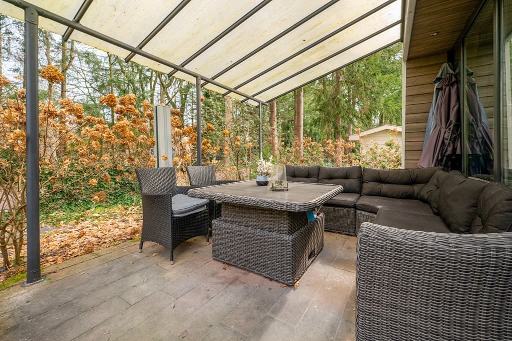 Overdekt terras met comfortabele zithoek en eettafel, omringd door groene natuur en herfstachtige begroeiing.
