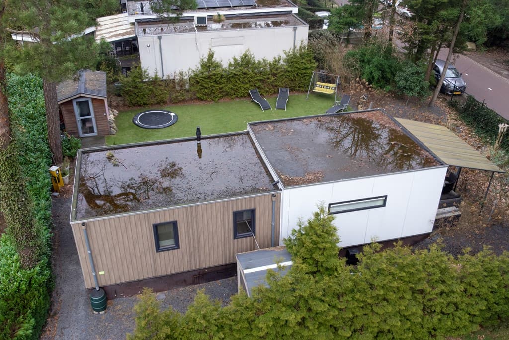 Recreatiewoning met plat dak en groen grasveld rondom, omgeven door bomen en enkele speeltoestellen.