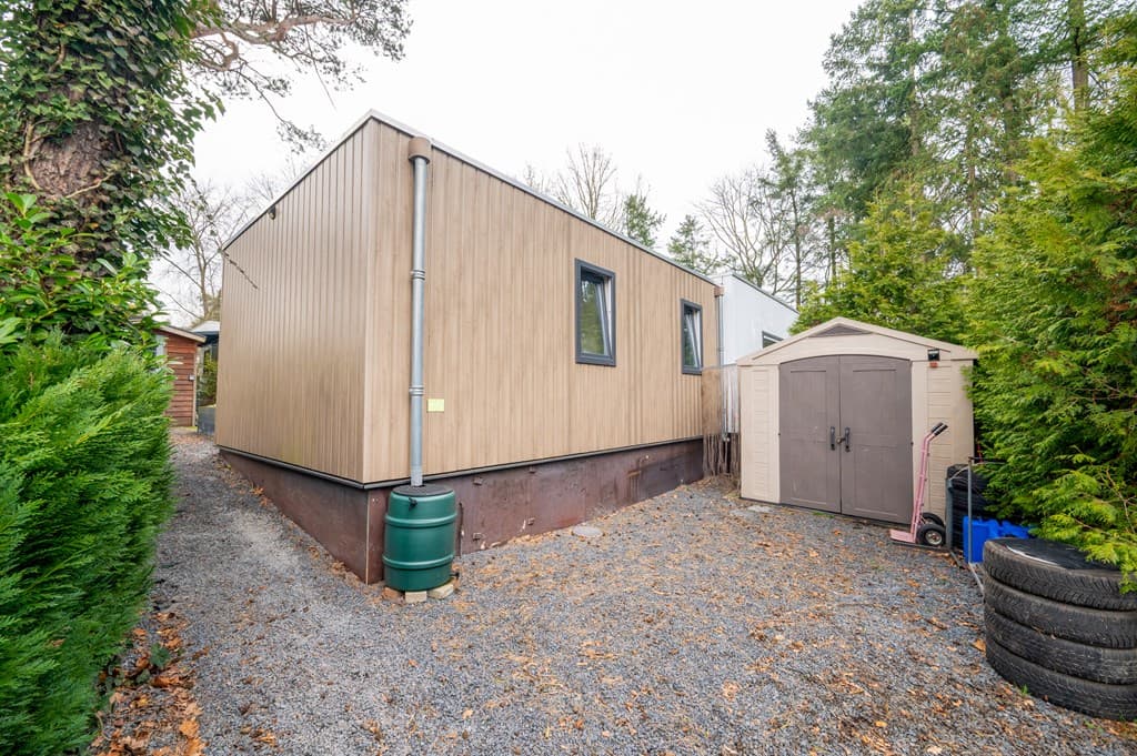 Zijaanzicht van een moderne recreatiewoning met houten gevel omgeven door groen en een berging op een grindpad