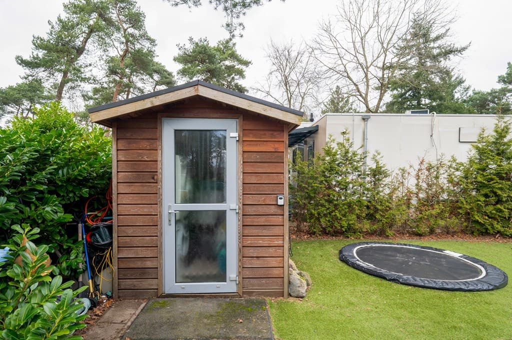 Houten tuinhuisje met glazen deur en trampoline op een grasveld, omringd door groene bomen en struiken.