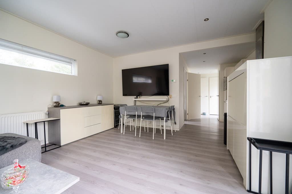 Lichte woonkamer met een eettafel en stoelen, een televisie aan de wand en openslaande deuren naar een hal.