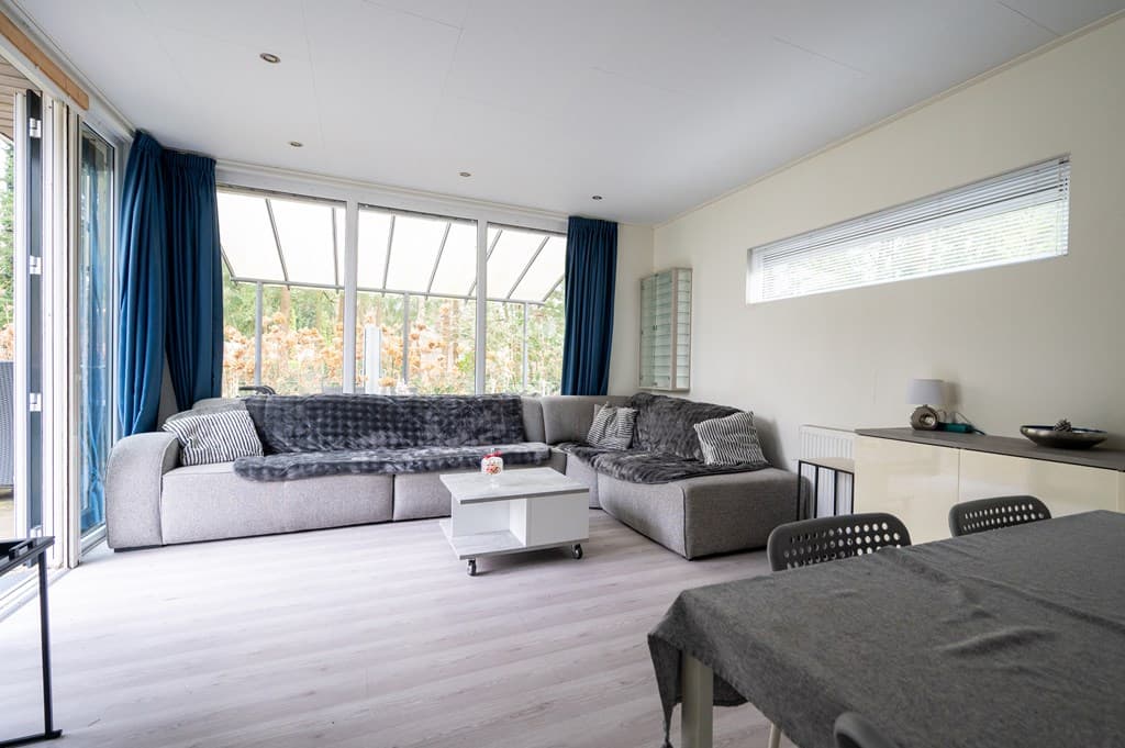Moderne woonkamer met grote raampartijen, lichte wanden en een hoekbank in grijstinten, vergezeld door een eettafel.