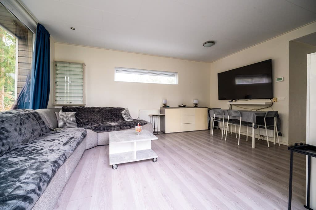 Woonkamer met een grote hoekbank, tv, salontafel en eettafel met stoelen bij een raam met uitzicht op de tuin
