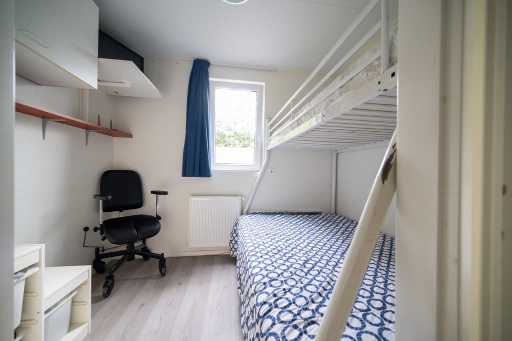 Kamertje met een stapelbed, bureau met stoel en raam met gordijn, lichte vloer en eenvoudige inrichting.