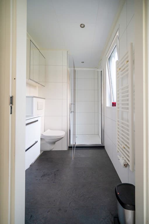 Moderne badkamer met douche, toilet, wastafel en radiator, witte tegels en een raam voor natuurlijk licht