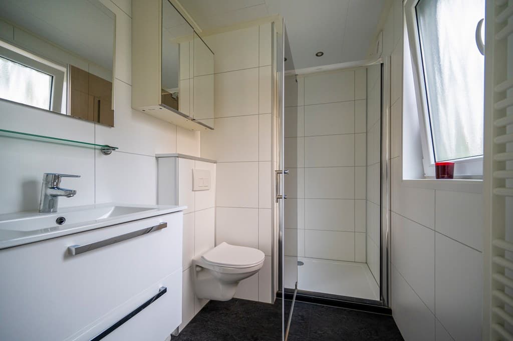 Moderne badkamer met douche, wastafel en toilet, witte wandtegels en grote ramen voor natuurlijk licht.