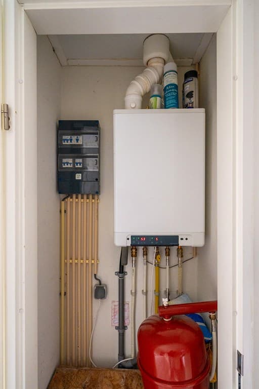 Technische ruimte met verwarmingsketel, leidingen en een rode expansievat in een schuur.