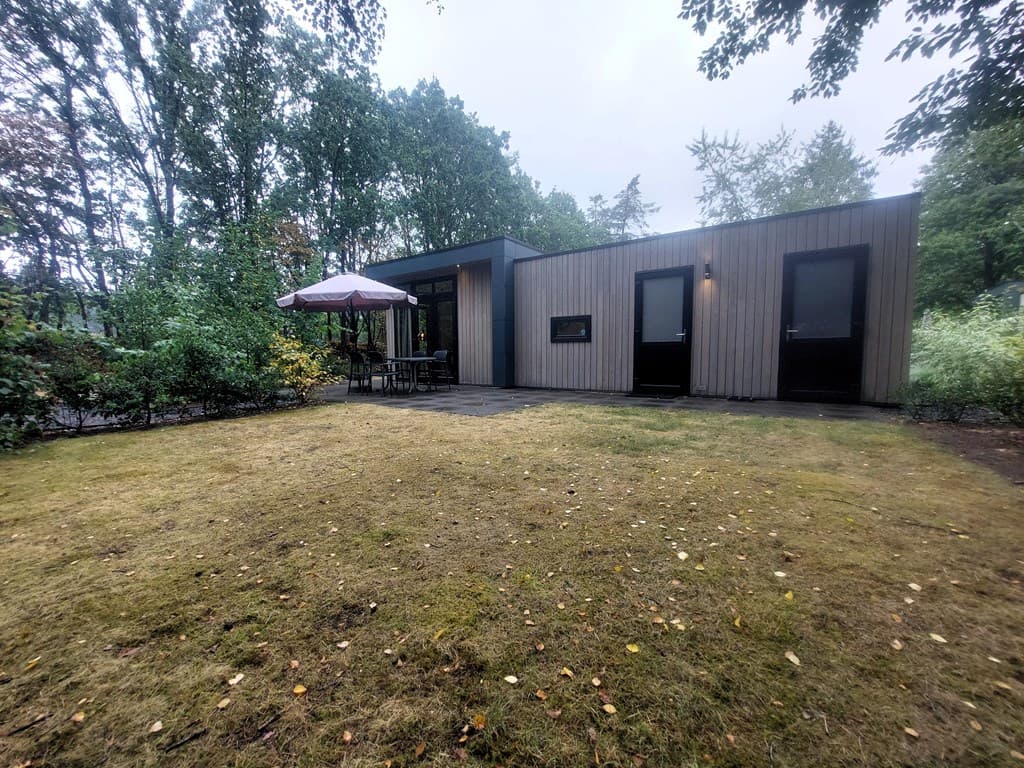 Moderne recreatiewoning met een houten gevel, tuin en terras onder een parasol, omgeven door bomen