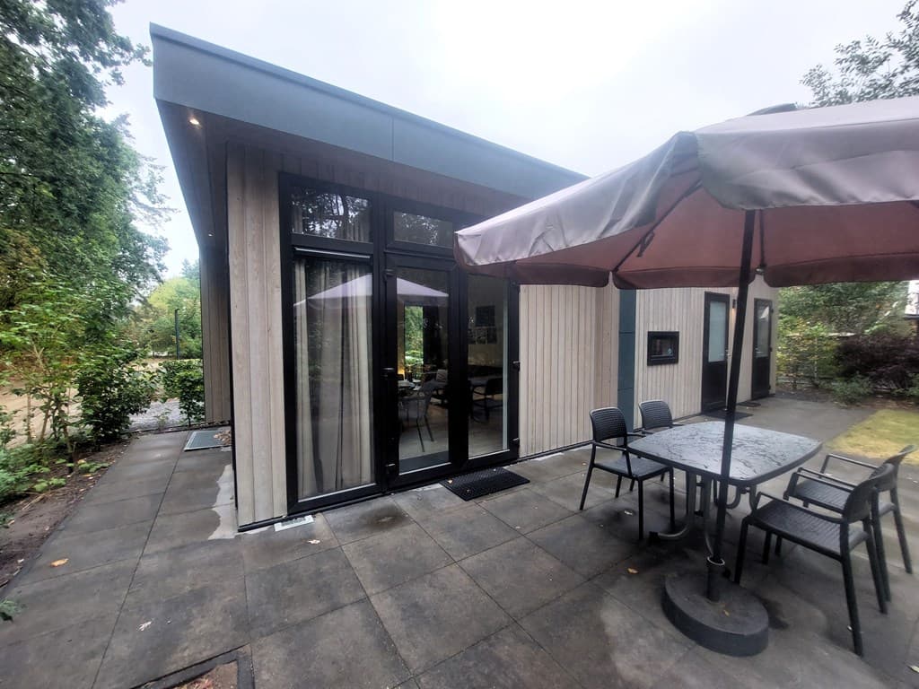 Moderne recreatiewoning met glazen schuifdeuren en een terras met parasol en tafel op een betegelde vloer.
