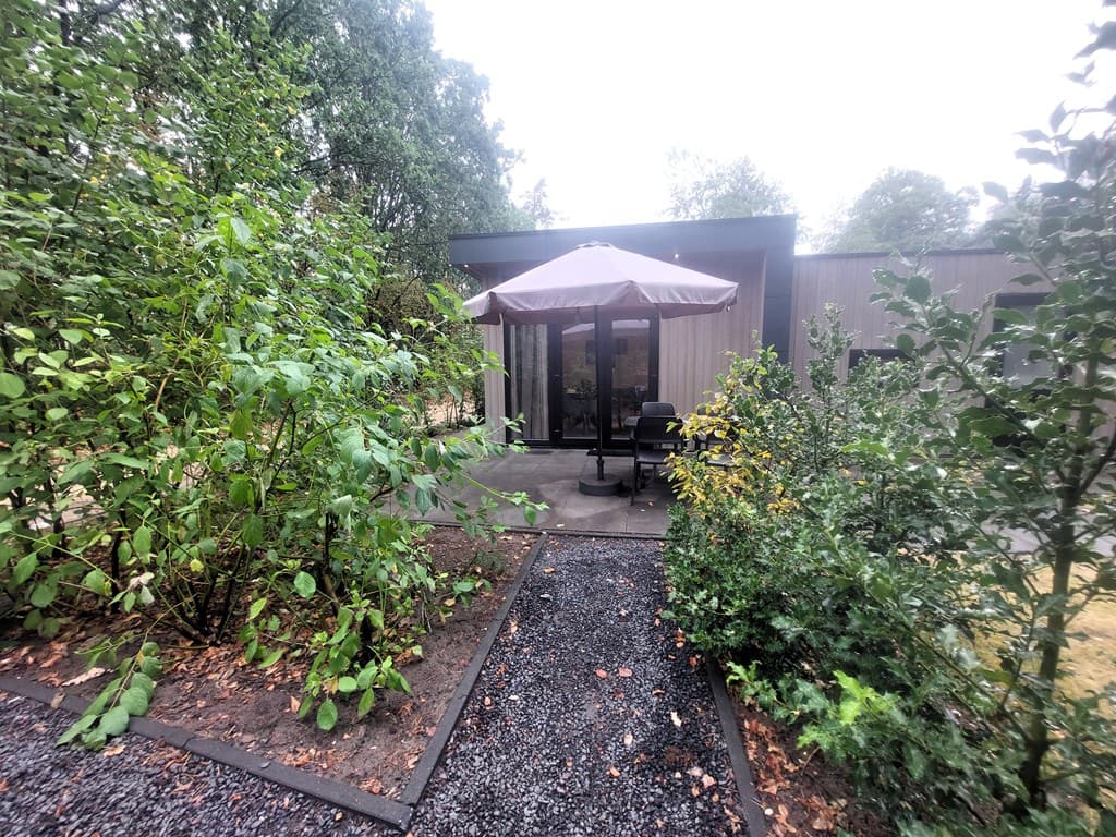 Vooraanzicht van een recreatiewoning met een terrras en een parasol, omgeven door groene planten en grindpaden