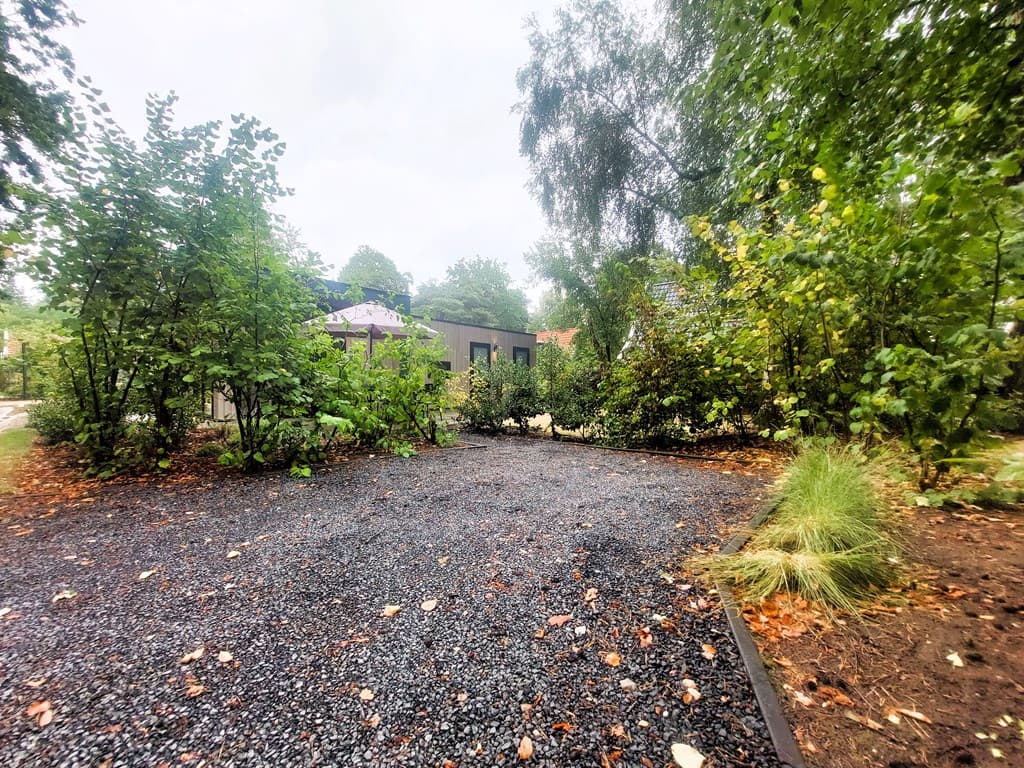 Recreatiewoning omringd door groene struiken en bomen, met een grindpad en een stukje gras zichtbaar.