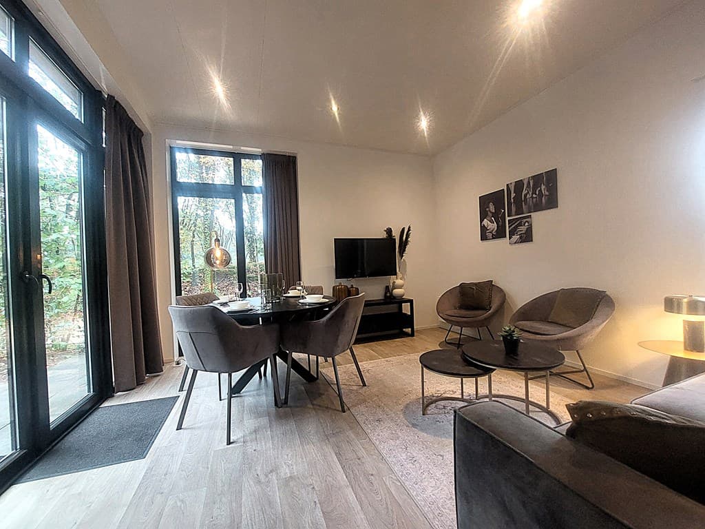 Moderne woonkamer met grote ramen, comfortabele stoelen, een tv en een eettafel met stoelen in een bosrijke omgeving