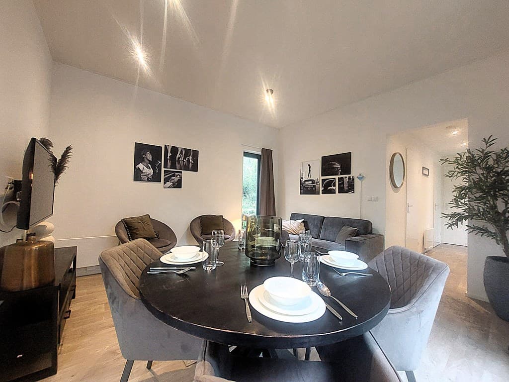 Eetkamer met een ronde tafel, stoelen, een televisie en foto’s aan de wand, uitzicht op natuur door een raam.
