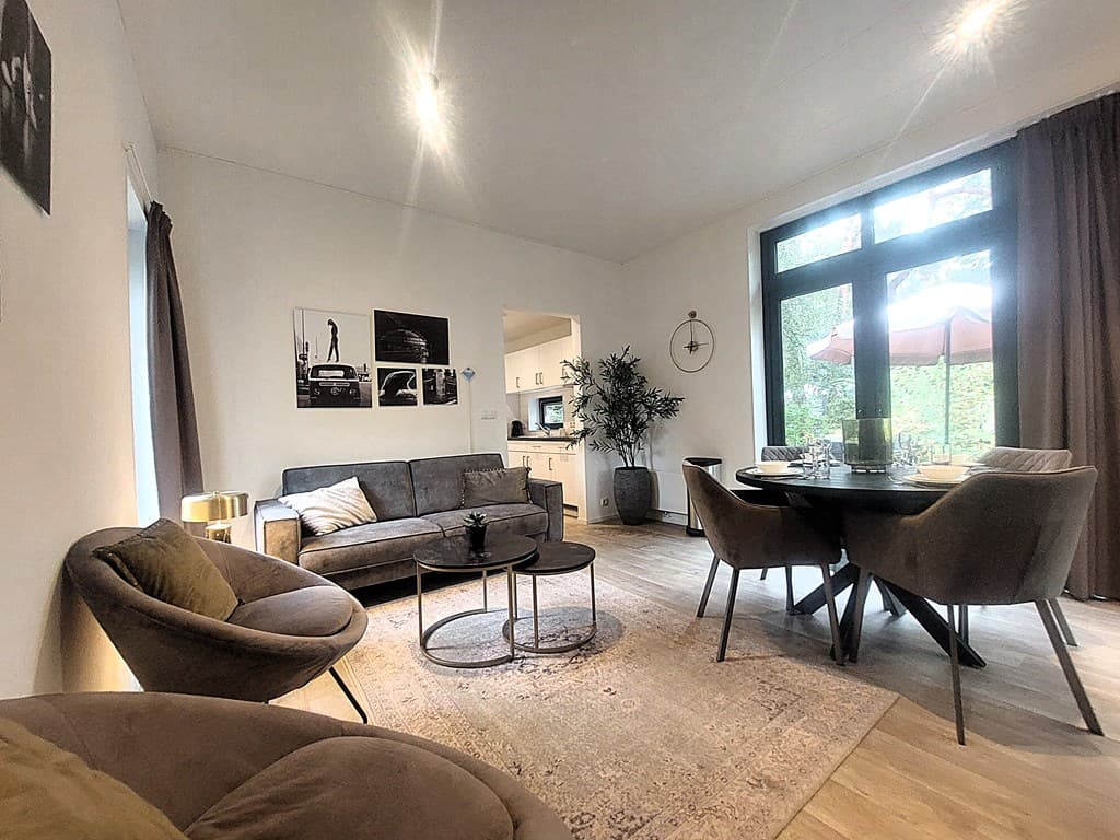 De woonkamer met een bank, twee stoelen en een eettafel, grote ramen met uitzicht op de buitenruimte, kunst aan de muur.