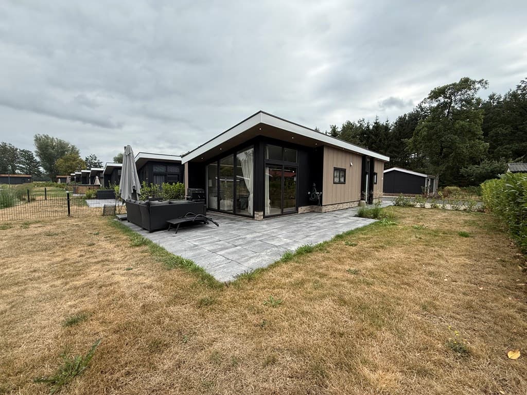 Moderne recreatiewoning met een glazen facade, een betegeld terras en groen gras rondom, omringd door bomen.