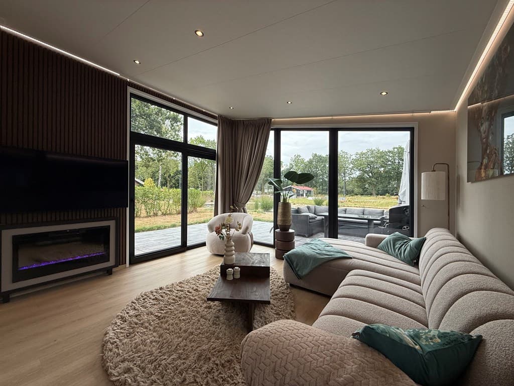 Lichte woonkamer met grote ramen, comfortabele bank, vloerkleed en zicht op het groene buitengebied.