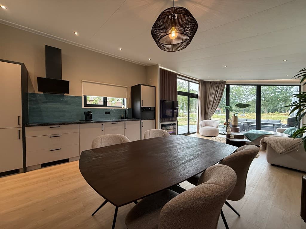 Moderne woonkamer met open keuken, grote ramen naar buiten en een houten eettafel met lichte stoelen.