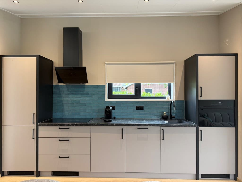 Moderne keuken met lichte kasten, zwart aanrechtblad en blauwe wandtegels, voorzien van kookplaat en inbouwapparatuur.