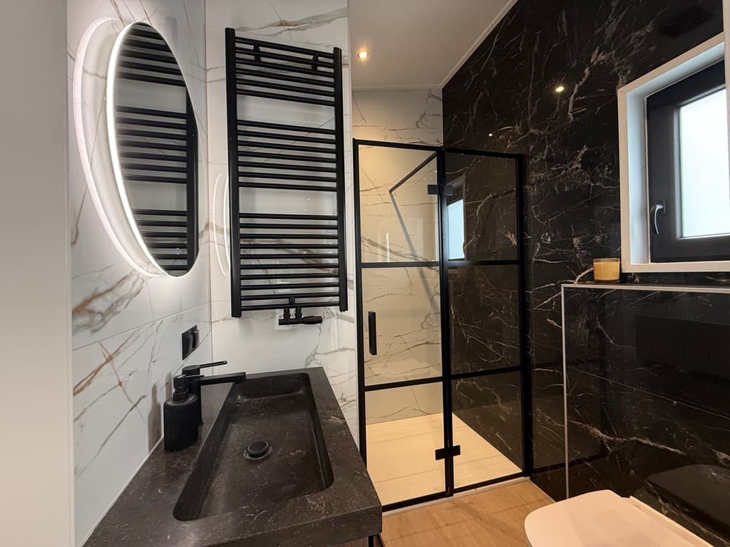 Moderne badkamer met zwarte marmerlook wand, inloopdouche met glazen wand en een ronde spiegel boven het wastafelmeubel.